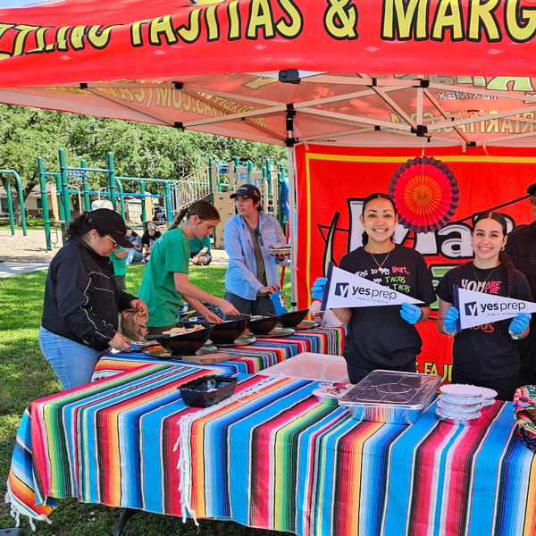 Maria Rita's Tex-Mex staff serving food under a red tent with a colorful striped tablecloth.
