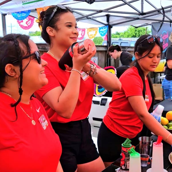 Three women in red shirts and sunglasses prepare drinks at an outdoor bar with colorful decorations.