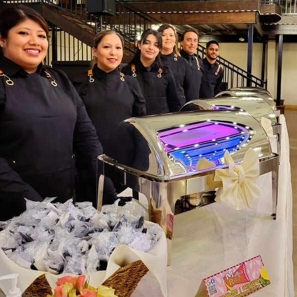 Maria Rita's staff smiling behind a buffet line with chafing dishes and individually wrapped utensils.