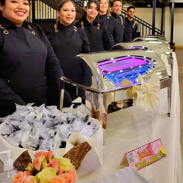 Maria Rita's staff smiling behind a buffet line with chafing dishes and individually wrapped utensils.