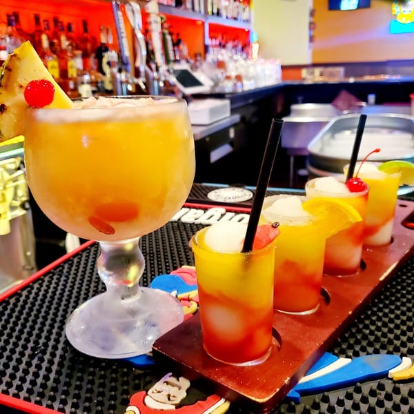 A large Tex-Mex cocktail with pineapple and cherry, next to a flight of four smaller colorful drinks.